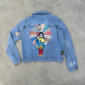 May include: A light blue denim jacket with a colorful patchwork design on the back. The design features a girl holding a cat, with a floral pattern surrounding them.