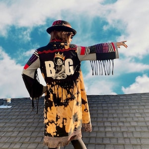 May include: A black and yellow tie-dye jacket with a black and white image of a young boy wearing a crown and the word "BIG" on the back. The jacket has fringe on the sleeves and bottom hem. The person wearing the jacket is standing on a roof with a blue sky and white clouds in the background.