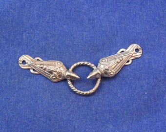 Puede incluir: Un broche de metal plateado para capa con dos terminales simétricos en forma de ave con grabados detallados, unidos por un anillo circular trenzado central. Cada terminal de ave incluye un pequeño orificio circular en el extremo.
