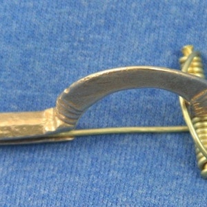 May include: A gold-colored metal brooch with a curved design and a spring clasp. The brooch has a decorative pattern on the curved part.