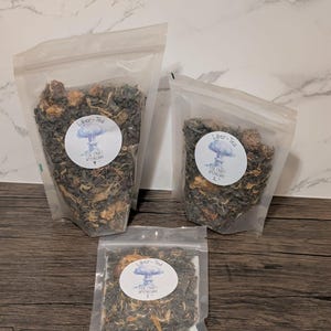 May include: Three clear plastic bags filled with loose leaf tea. Each bag has a white label with the text "Liber-Tea" and a graphic of a mushroom cloud. The tea appears to be a mix of green and brown leaves with some visible dried fruit.