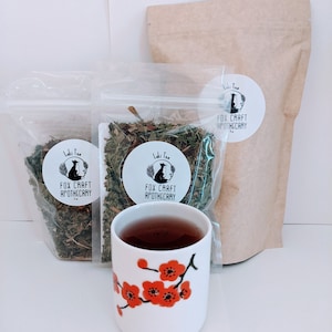 May include: Two clear plastic bags filled with dried herbs, a brown paper bag with a white label that says "Loki Tea Fox Craft Apothecary", and a white teacup with red cherry blossoms on it. The teacup is filled with dark tea.