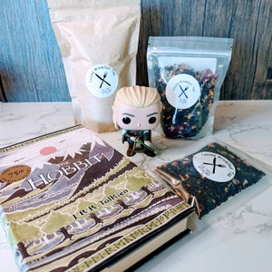 May include: A brown paper bag with the text "Second Breakfast Tea" printed on it, a bag of loose leaf tea with the text "Second Breakfast Tea" printed on it, a book titled "The Hobbit" by J.R.R. Tolkien, and a small plastic figure of a character from the Lord of the Rings.