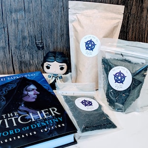 May include: A book titled "The Witcher: Sword of Destiny Illustrated Edition" by Andrzej Sapkowski, a Funko Pop figure of a woman with brown hair, and three bags of tea labeled "The Last Wish Tea for Craft Witchery".
