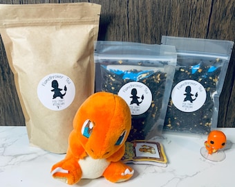 May include: A brown paper bag with a white label that says "Flamethrower Tea" and a black silhouette of a Pokemon character. Two clear plastic bags with black tea leaves and white labels that say "Flamethrower Tea" and a black silhouette of a Pokemon character. An orange plush Pokemon character and a small orange plastic Pokemon figurine.