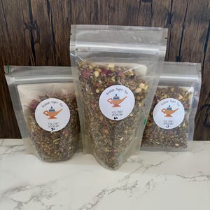 May include: Three clear plastic bags filled with loose leaf tea. Each bag has a circular label with a teapot illustration and the text "Arabian Night Tea" and "Fox Craft Apothecary". The bags are on a marble surface.