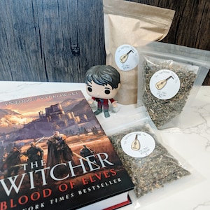 May include: A brown book titled "The Witcher: Blood of Elves" by Andrzej Sapkowski, a brown paper bag with a sticker that says "The Nightingale Prince Tea", a small figurine of a boy with brown hair wearing a red shirt and blue jeans, and two clear plastic bags filled with loose leaf tea with stickers that say "The Nightingale Prince Tea" and a picture of a lute.