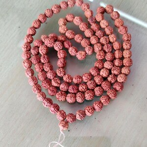 Può includere: Una collana di perline marrone-rossastre e strutturate. Le perline sono infilate con un filo bianco, formando più fili. Le perline hanno un aspetto ruvido e organico. La collana è appoggiata su una superficie chiara.