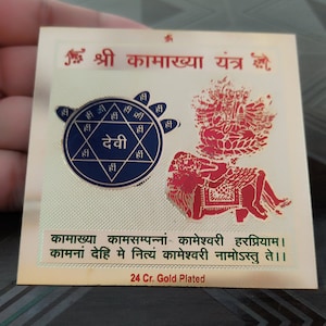 Puede incluir: Una placa de metal chapada en oro con una imagen roja y azul de una deidad. La placa presenta el texto "श्री कामाख्या यंत्र" y "कामाख्या कामसम्पन्नां कामेश्वरी हरप्रियाम। कामनां देहि मे नित्यं कामेश्वरी नामोऽस्तु ते।।" y "24 Cr. Gold Plated".