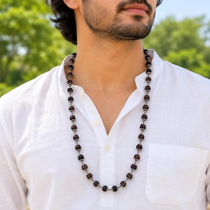 May include: A long necklace featuring dark brown, textured, spherical rudraksha beads separated by small, polished silver-toned metal spacers. The beads are strung in a continuous pattern, resting against a plain white button-down shirt.