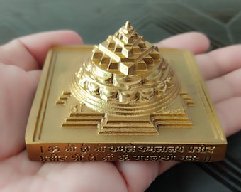 May include: A gold-colored metal pyramid-shaped object with intricate details and a Sanskrit inscription on the base. The inscription reads: "Om Shri Hi Shri Kamale Kamalalaye Prasid Prasid Shri Hi Shri Om Mahalaxmi Namah."