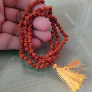 May include: A strand of prayer beads made of small, round, reddish-brown beads. The beads are strung together with orange thread and have a yellow tassel at the end. The beads are held in a person's hand.