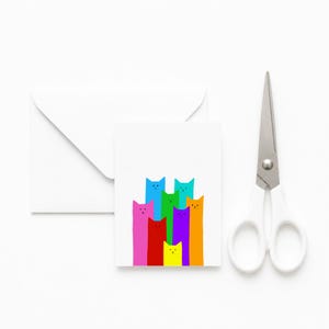 Puede incluir: Una tarjeta de felicitación blanca con una ilustración colorida de ocho gatos estilizados en rosa, rojo, amarillo, verde, azul, morado y naranja. La tarjeta viene acompañada de un sobre blanco y unas tijeras de metal plateado con mangos blancos.