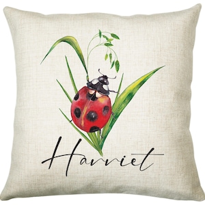 May include: A white linen cushion featuring a red and black ladybug on a green stem with the name "Harriet" printed in black.