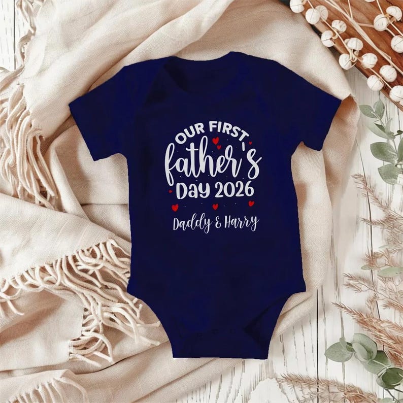 Op de afbeelding: Marineblau baby-rompertje met de tekst "OUR FIRST father's DAY 2026 Daddy & Harry" in witte letters. Rode hartjes accentueren het ontwerp. Het rompertje ligt op een cr&egrave;mekleurige deken.