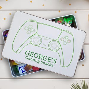 May include: A white rectangular tin featuring a green game controller outline and the text "GEORGE'S Gaming Snacks". The tin is open, displaying sweets. The scene is decorated with festive elements, including a bauble and pine cone.
