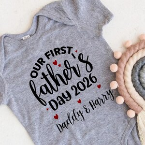 Op de afbeelding: Een grijze baby-onesie met de tekst "Our First Father's Day 2026 Daddy & Harry" in zwart script. Rode hartaccenten sieren de tekst. Een decoratieve regenboog is zichtbaar op de achtergrond.