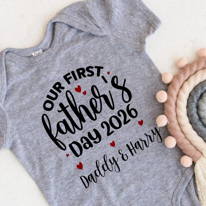 Op de afbeelding: Een grijze baby-onesie met de tekst "Our First Father's Day 2026 Daddy & Harry" in zwart script. Rode hartaccenten sieren de tekst. Een decoratieve regenboog is zichtbaar op de achtergrond.