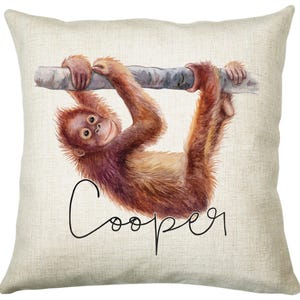 May include: A beige throw pillow featuring a watercolour illustration of an orangutan hanging from a branch. The orangutan is brown and orange with a light brown face. The text "Cooper" is written in black cursive below the orangutan.