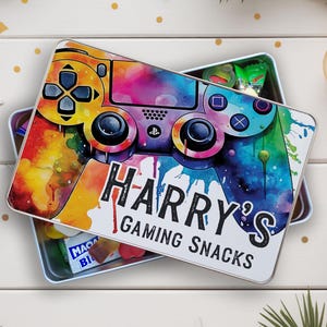 May include: A rectangular metal tin featuring a colourful watercolour design of a games controller. The tin is labelled "HARRY'S GAMING SNACKS" in black text. The tin is open, revealing snacks inside.