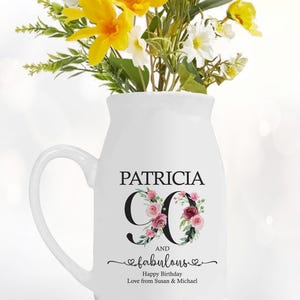 May include: White ceramic milk jug with a handle, decorated with pink and green floral designs and the text "Patricia and fabulous" with a heart. The number 90 is in black with a floral design around it. The text "Happy Birthday Love from Susan & Michael" is also included.
