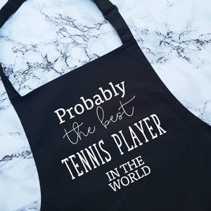 May include: Black apron with white text that reads "Probably the best tennis player in the world".