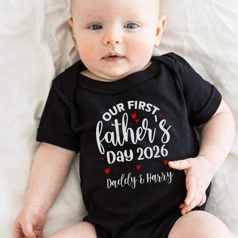 Op de afbeelding: Een zwarte babyromper met de tekst "OUR FIRST Father's Day 2026 Daddy & Harry" in witte letters en rode hartjes. De baby ligt op een witte ondergrond.