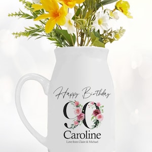 May include: White ceramic milk jug with a handle, decorated with pink and green floral designs and the text "Happy Birthday 90 Caroline Love from Claire & Michael".