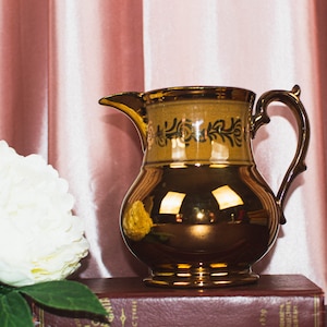 May include: A gold-colored pitcher with a decorative handle and spout. The pitcher has a bulbous body and a band of dark green floral design near the top. A white flower and a book are in the foreground. The background is a pink curtain.