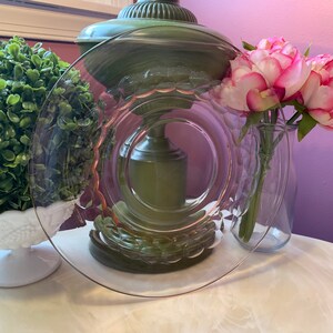 May include: A clear glass plate with a decorative rim, resting on a white surface. The plate is next to a vase of pink and white flowers. A green lamp and a white decorative bowl are also in the background.