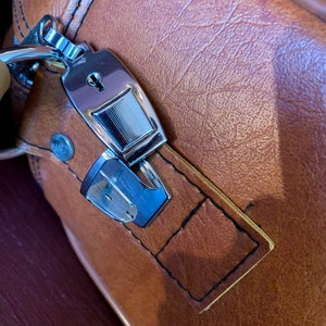 May include: A brown leather suitcase with a silver lock and key. The lock has a keyhole and a small metal plate with a design on it.