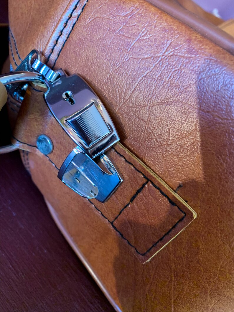 May include: A brown leather suitcase with a silver lock and key. The lock has a keyhole and a small metal plate with a design on it.