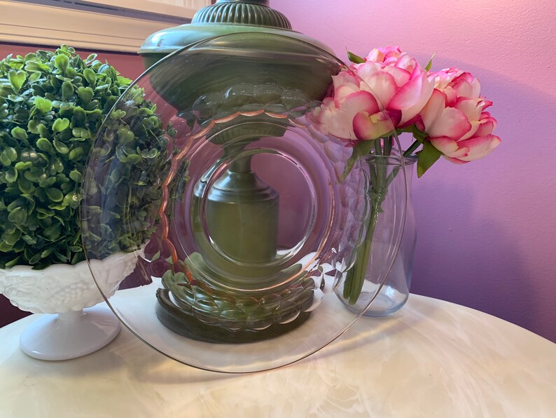 May include: A clear glass plate with a decorative rim, resting on a white surface. The plate is next to a vase of pink and white flowers. A green lamp and a white decorative bowl are also in the background.