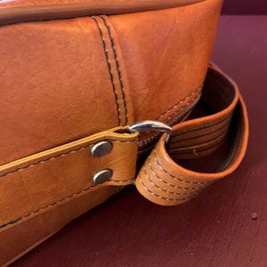 May include: Close-up of a brown leather bag with a silver metal buckle and a black strap. The bag has a stitched design and a flap closure.