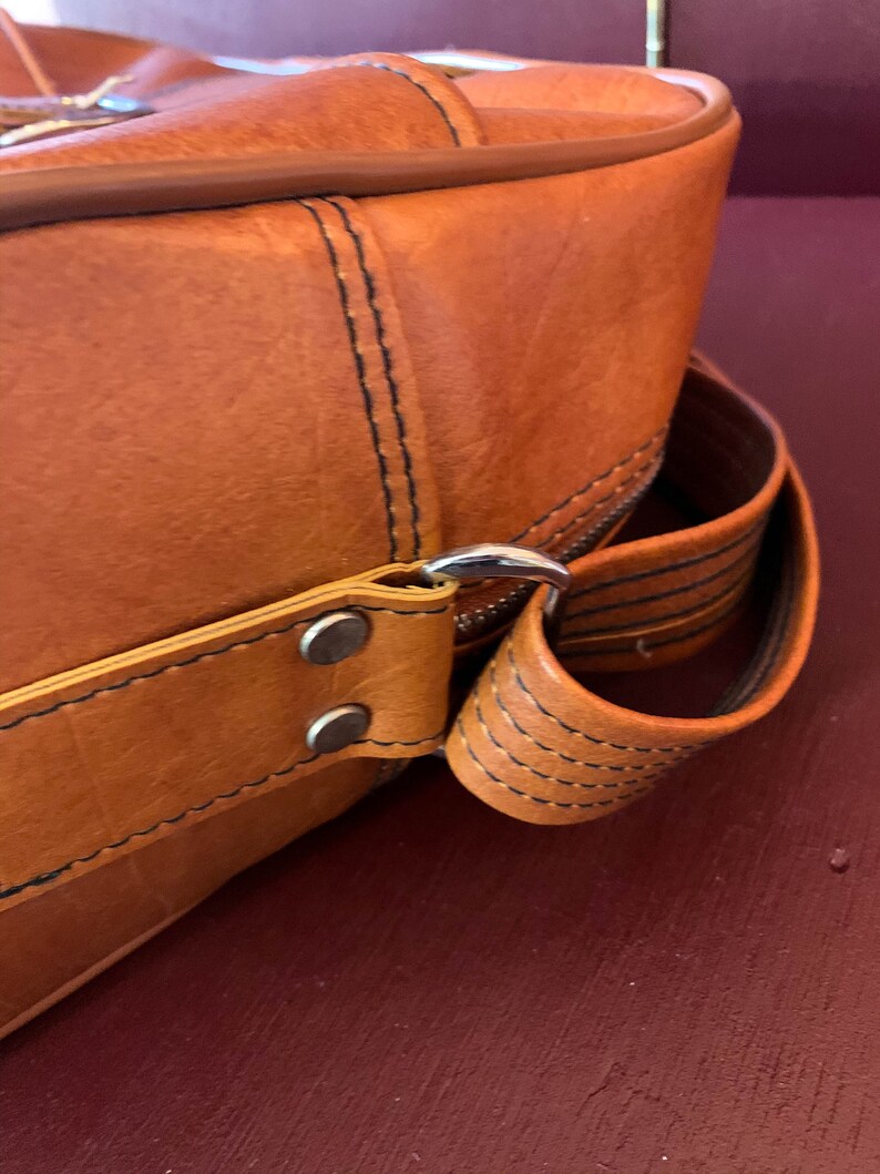 May include: Close-up of a brown leather bag with a silver metal buckle and a black strap. The bag has a stitched design and a flap closure.