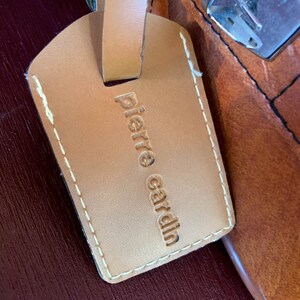 May include: A brown leather luggage tag with white stitching and the text "Pierre Cardin" embossed in gold.