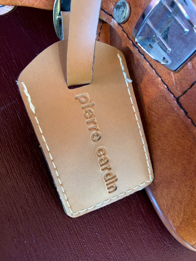 May include: A brown leather luggage tag with white stitching and the text "Pierre Cardin" embossed in gold.