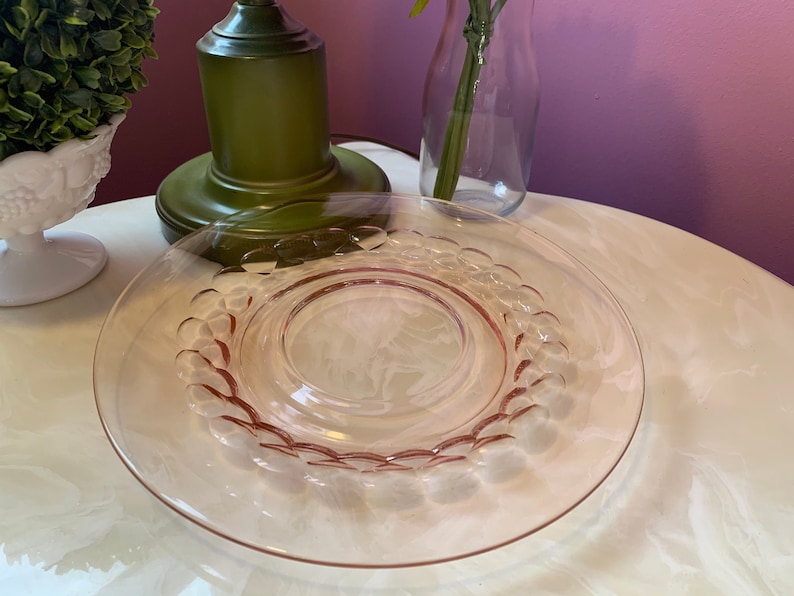 May include: A clear, pink-tinted glass plate with a decorative scalloped edge. The plate has a circular design in the center and is set on a white surface. A green lamp and a small white decorative object are in the background.