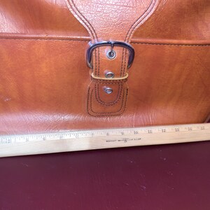 May include: A brown leather satchel with a silver buckle. The bag is sitting on a wooden ruler.