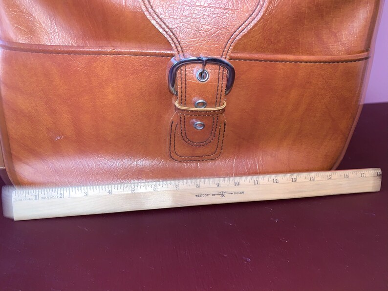 May include: A brown leather satchel with a silver buckle. The bag is sitting on a wooden ruler.