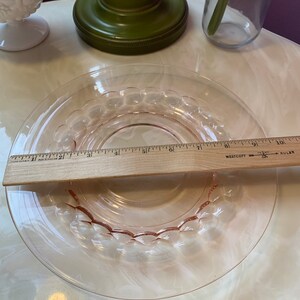 May include: A clear, pink glass plate with a decorative bubble design around the rim. A wooden ruler is placed across the plate, measuring approximately 6 inches in diameter. The plate is on a white surface.