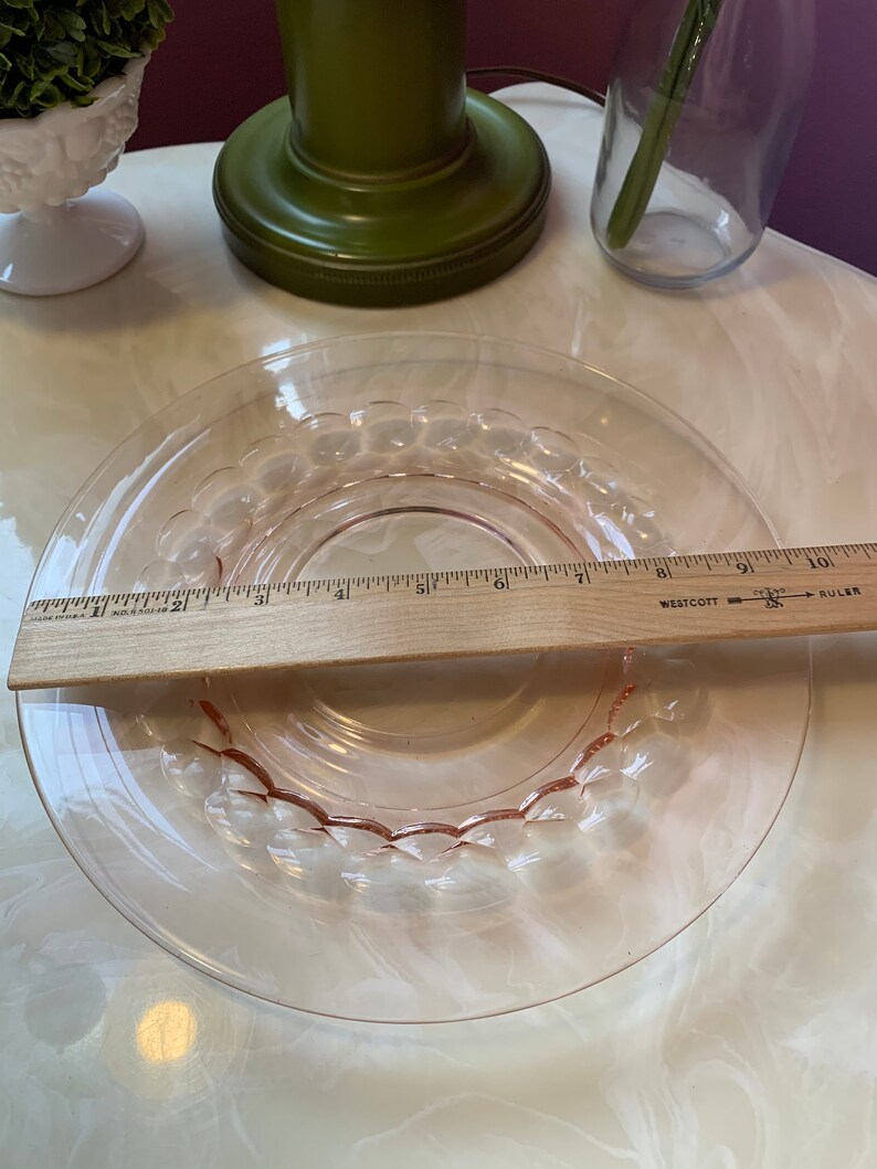 May include: A clear, pink glass plate with a decorative bubble design around the rim. A wooden ruler is placed across the plate, measuring approximately 6 inches in diameter. The plate is on a white surface.