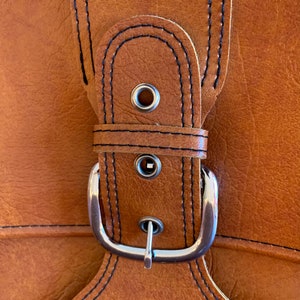 May include: Close-up of a brown leather strap with a silver buckle. The strap has black stitching and two metal eyelets.
