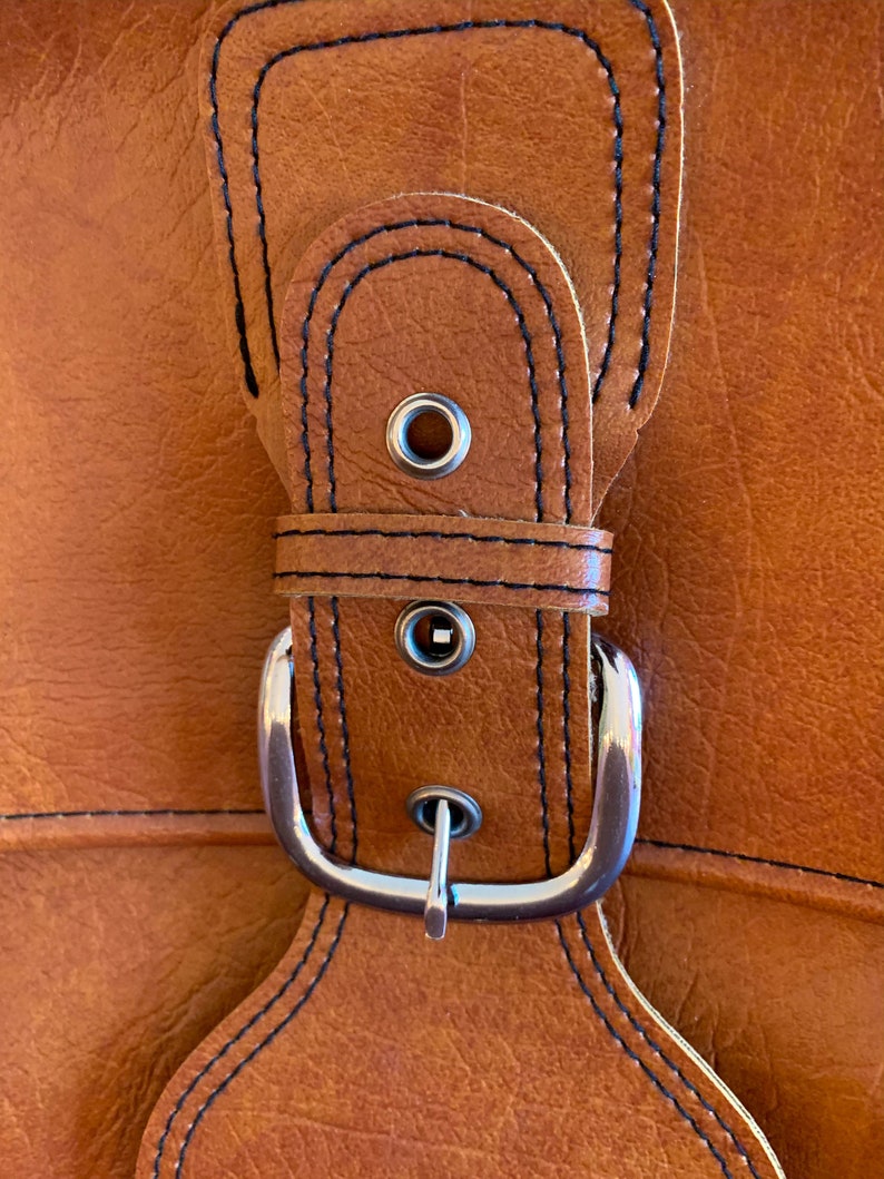 May include: Close-up of a brown leather strap with a silver buckle. The strap has black stitching and two metal eyelets.