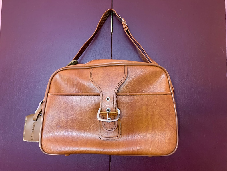 May include: A brown leather travel bag with a buckle closure and a small leather tag that says "Pierre Cardin".