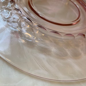 May include: A clear, pink glass plate with a decorative, textured rim. The plate has a smooth, oval-shaped center and a slightly raised edge. The rim features a repeating pattern of raised, rounded shapes. The plate is likely vintage glassware.