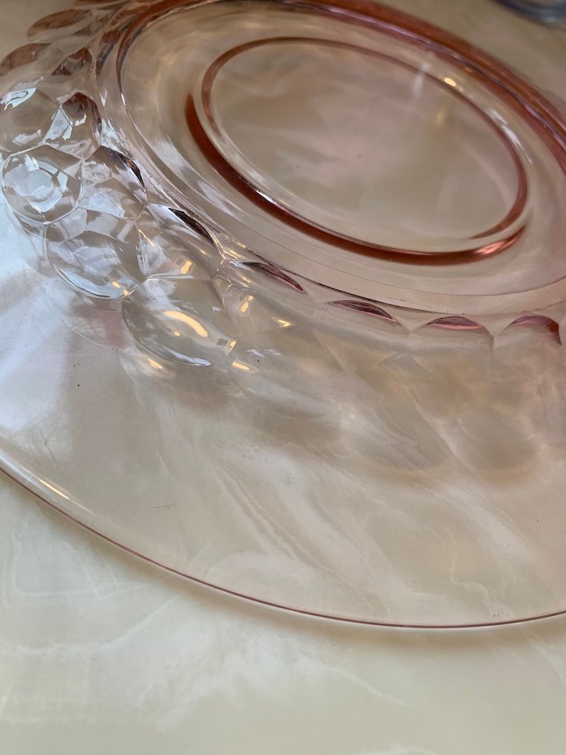 May include: A clear, pink glass plate with a decorative, textured rim. The plate has a smooth, oval-shaped center and a slightly raised edge. The rim features a repeating pattern of raised, rounded shapes. The plate is likely vintage glassware.