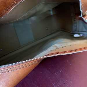 May include: The inside of a brown leather travel bag with a zipper closure. The bag is lined with a beige fabric.