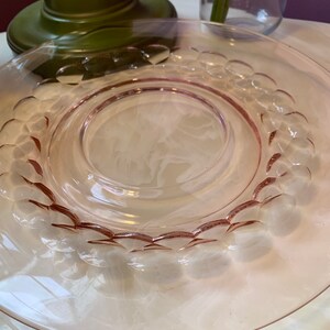 May include: A clear pink glass plate with a decorative bubble pattern around the rim. The plate has a circular design in the center and is set on a white surface. The plate is part of a dinnerware set.