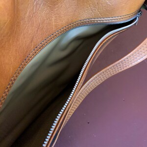 May include: A brown leather bag with a zipper closure and a brown leather strap. The bag is open and the inside is lined with a beige fabric.